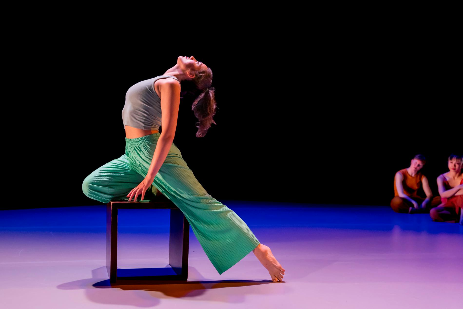 Alex, a dancer in all green, perches on a box and arches her back to look upward while other dancers look on.