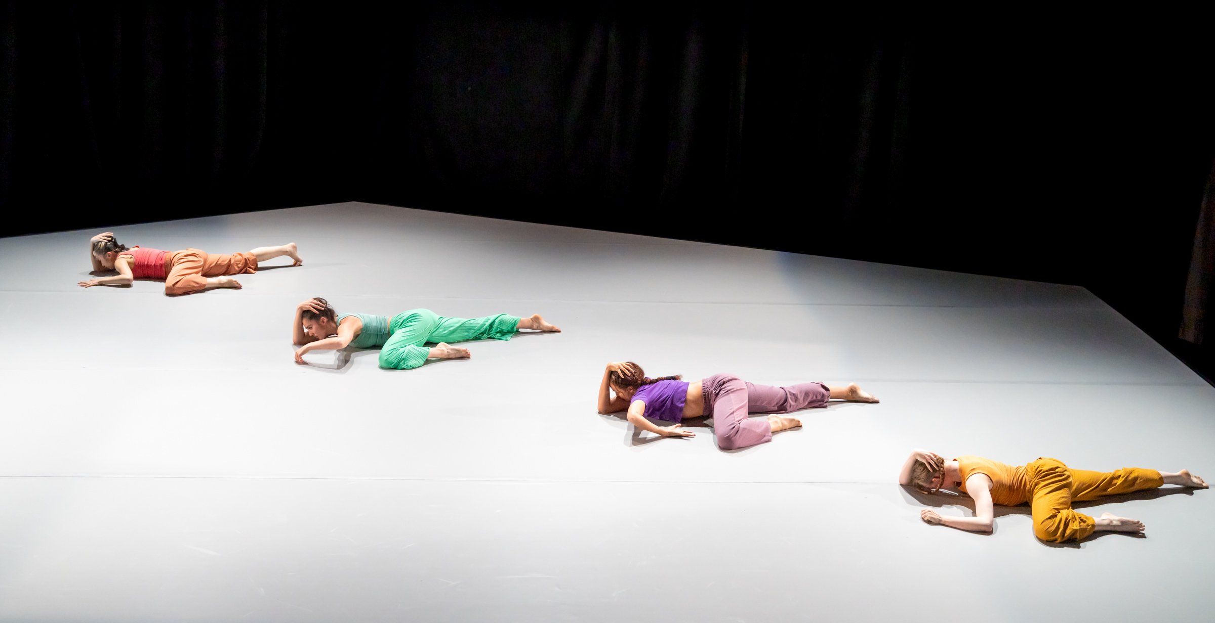 Four dancers, each wearing a different color, lay in nearly identical positions on a stark floor.